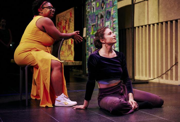 A white woman sits straight on a floor stage next to a Black woman sitting in a chair.